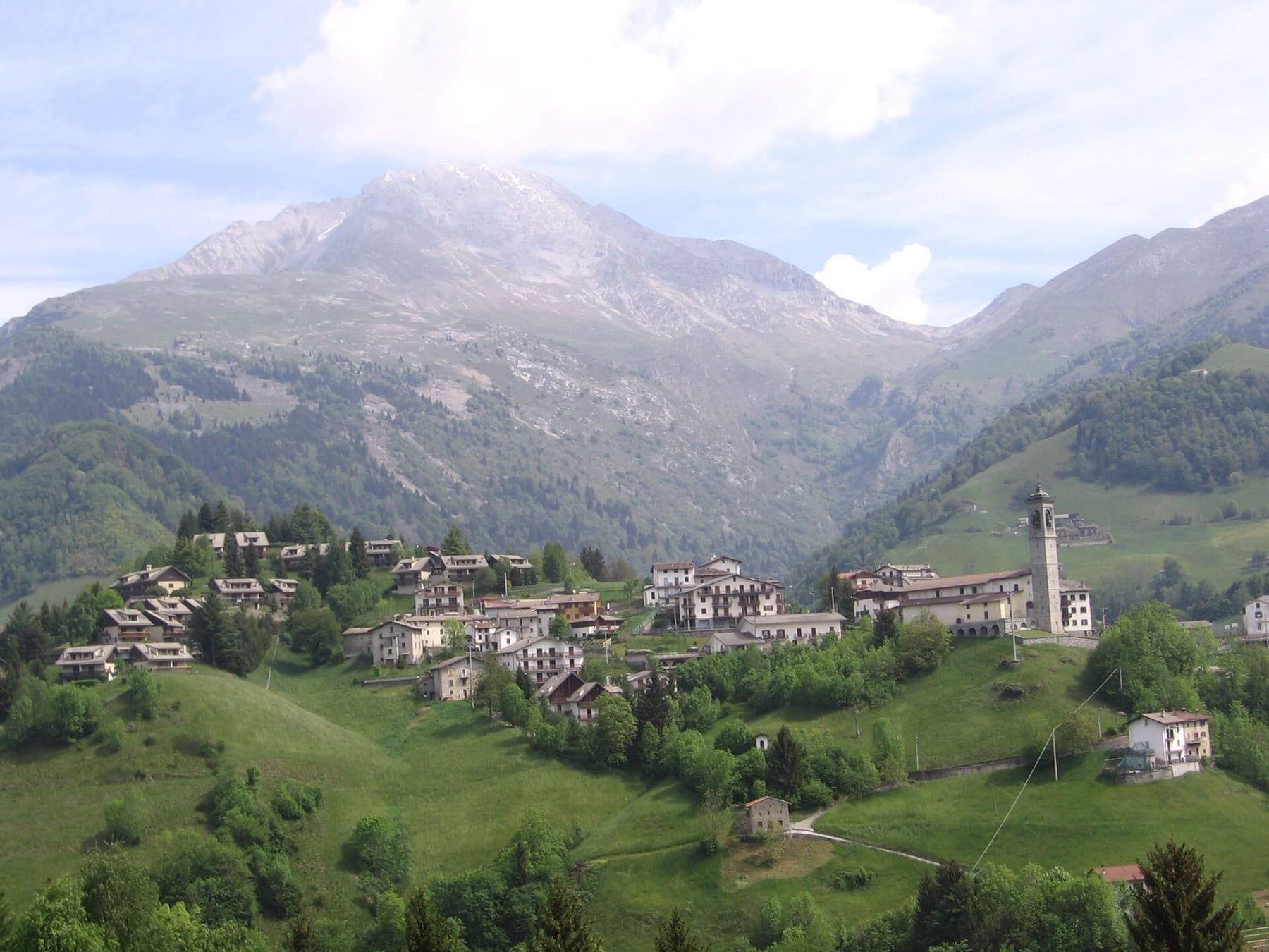 Zambla Bassa, frazione di Oltre il Colle, con il campanile e le case sui prati verdi ai piedi delle cime rocciose